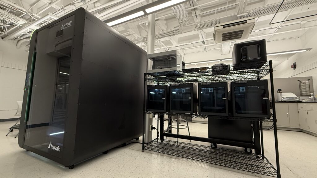The AMIC laboratory featuring a large, black Mosaic Array automated 3D printing system alongside a wire shelving unit holding several smaller desktop 3D printers. The room has high industrial ceilings with exposed piping and bright fluorescent lighting.