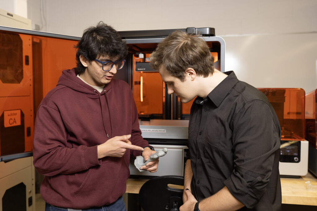 Students Frederic and Neel in the AMIC workshop examining a small, grey 3D-printed figurine. They are standing in front of several industrial 3D printers with orange protective covers.