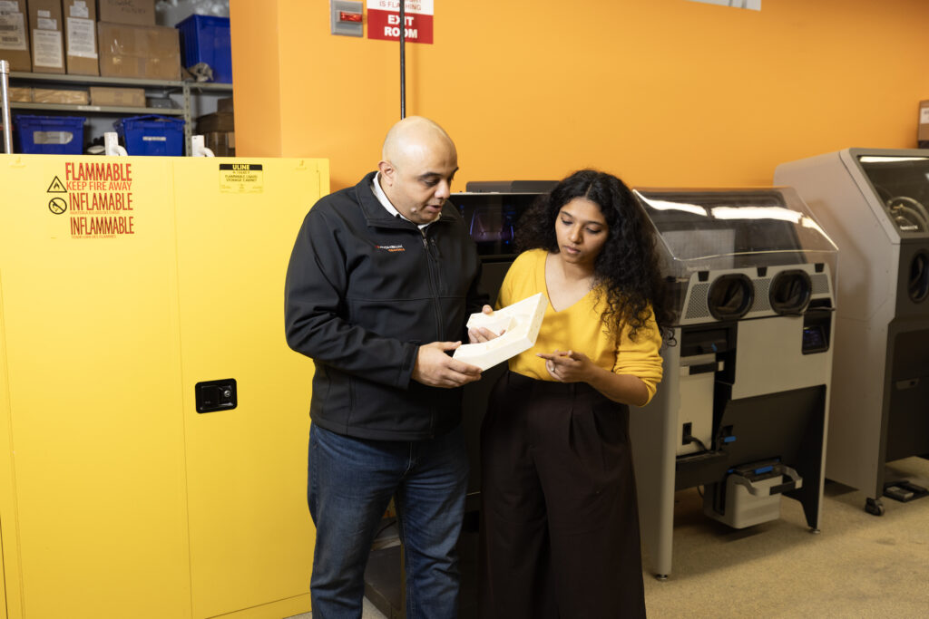 Researchers Sherif and Shrishti in the AMIC lab setting discussing a white, geometric 3D-printed part. In the background, there is a large yellow flammable storage cabinet and specialized post-processing equipment.