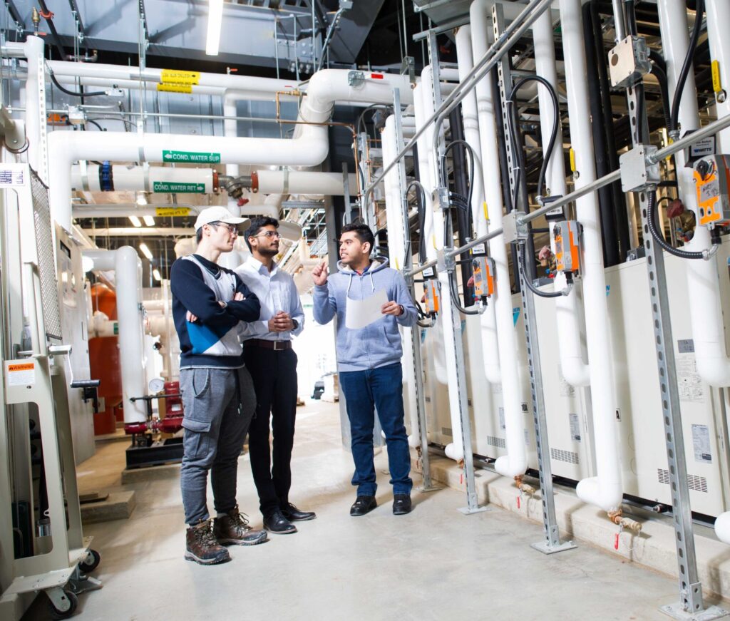 A diverse group of researchers in discussion in front of a series of pipes at Mohawk College