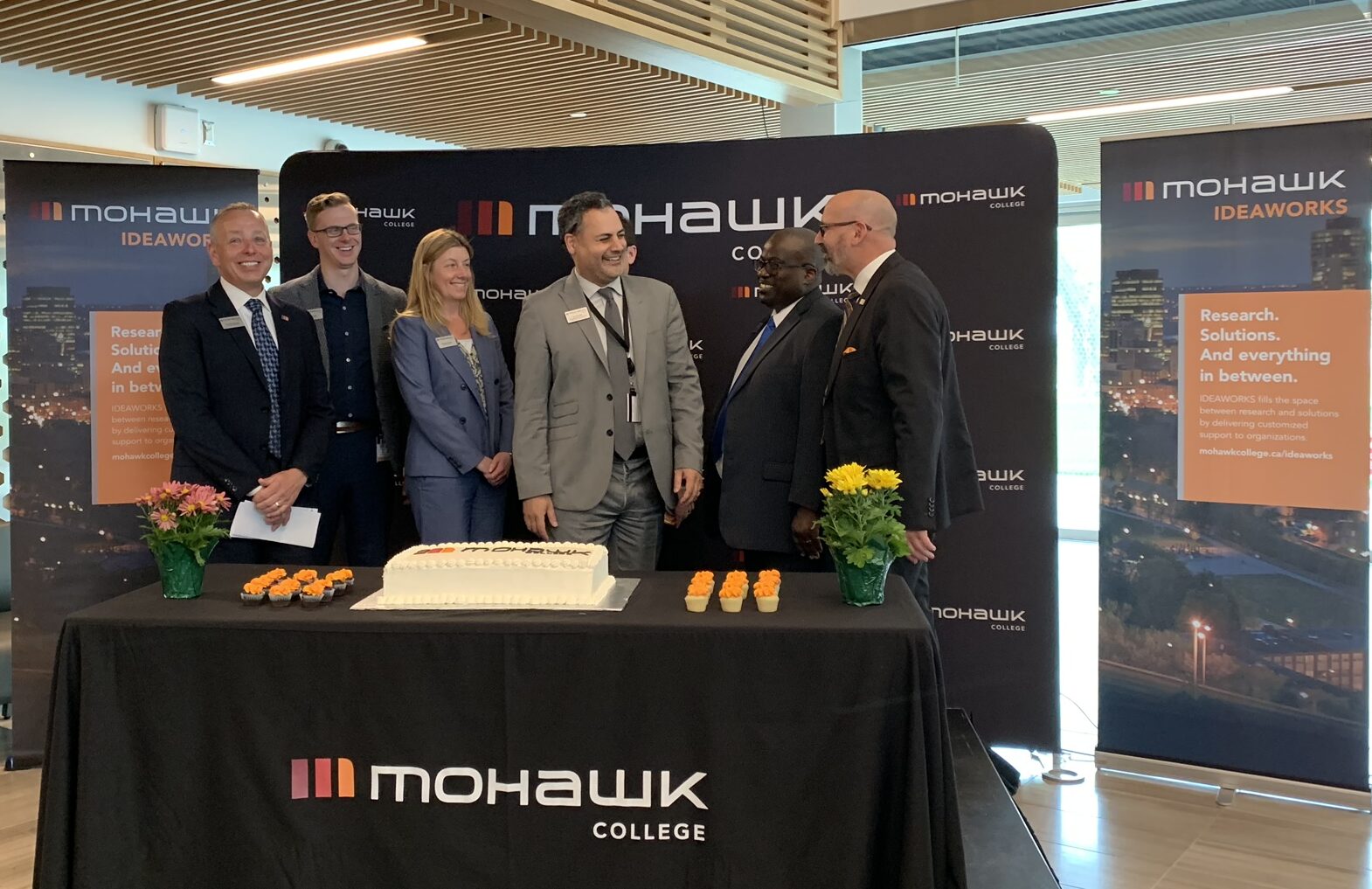 A diverse group of professionals standing behind a Mohawk College branded table, in the Joyce Center for Partnership and Innovation.